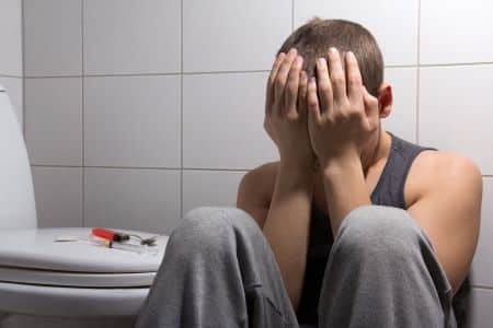 Man addicted to heroin sitting in a bathroom with drug supplies on the toilet