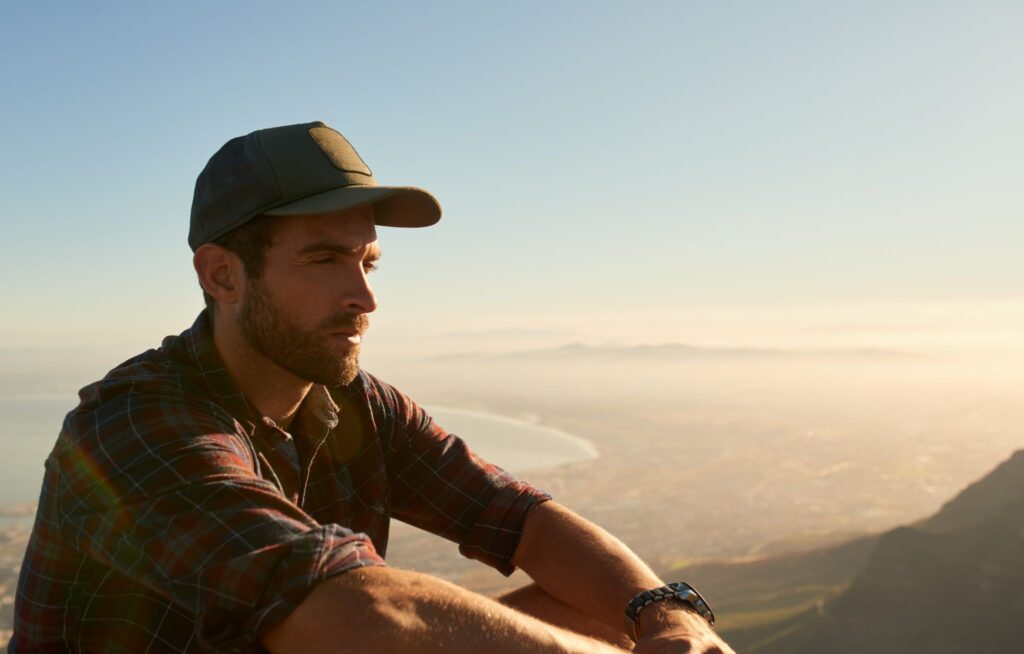 Man with a ball cap on sitting on a mountain overlooking a lake