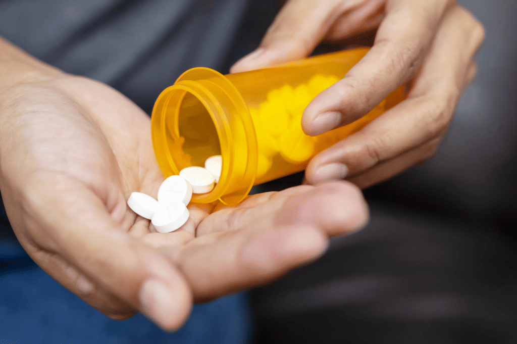 Close up of a person's hand holding a prescription bottle tipped over with white pills in hand, representing potential signs of opioid addiction that may require attention and support.