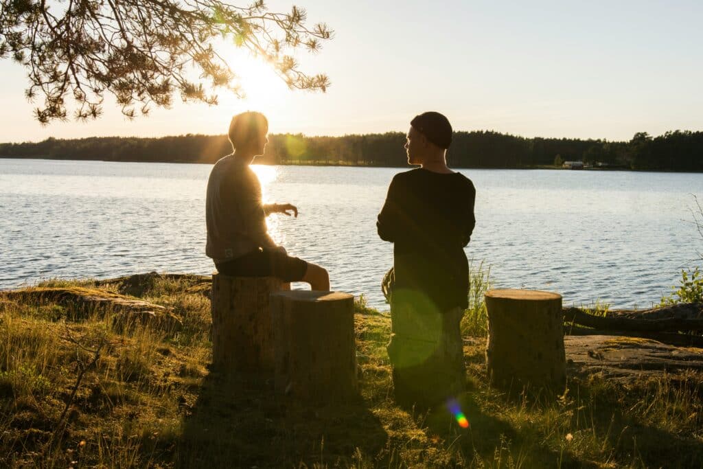 Two people sitting alongside a serene lakeside during sunset, engaged in a conversation and embodying person first language principles in their interaction.