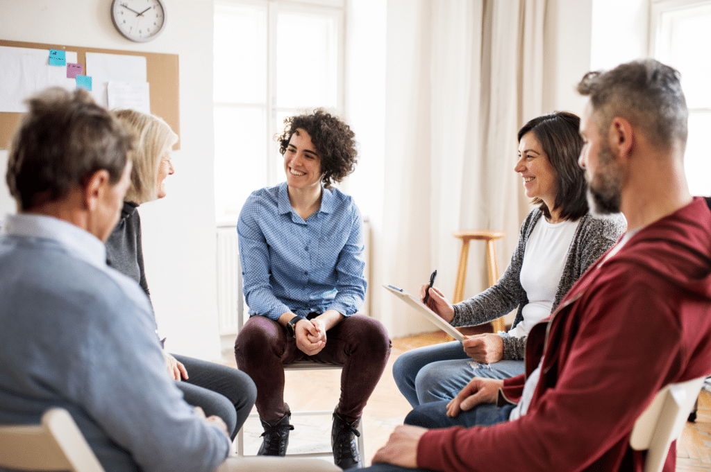 A group of individuals sitting in a circle at a counseling session, smiling and listening to a speaker, embodying support and understanding in the journey towards reducing the stigma of addiction for stigma reduction.